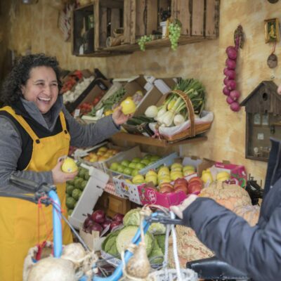 Photo by Rossella Fasoli Two women smiling and exchanging fresh produce at a vibrant greengrocer's market.