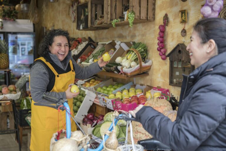 Two women smiling and exchanging fresh produce at a vibrant greengrocer's market.